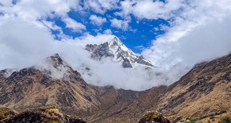 Pico andino cubierto de nieve se alza sobre montañas marrones escarpadas con cielo azul brillante y nubes dispersas.