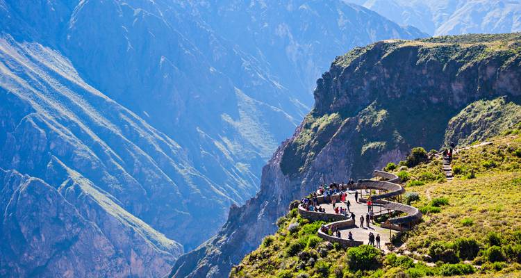 Mirador en el borde de un acantilado curvado lleno de visitantes con vista a un profundo cañón verde bajo cielos despejados.
