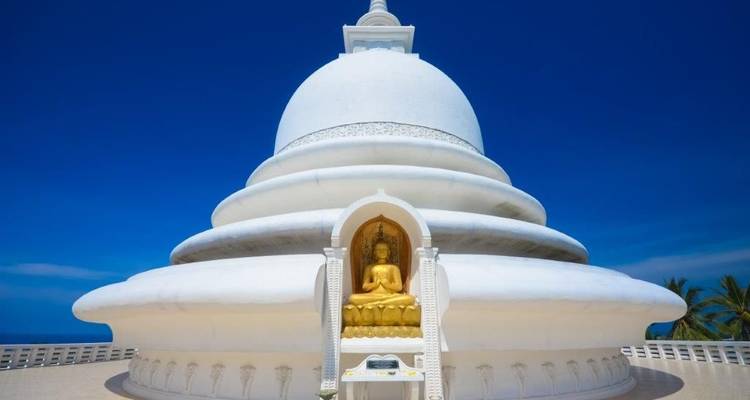 Briljant witte stupa aan de kust die oprijst tegen een levendige azuurblauwe hemel, met een gouden Boeddha in een nis.