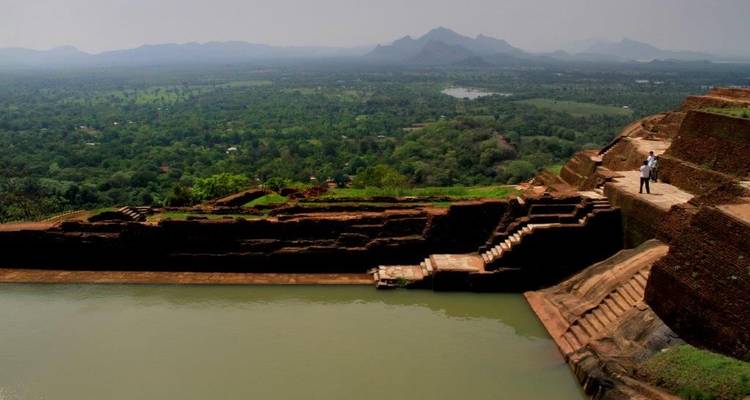 Uitzicht vanaf oude stenen ruïnes bovenop een plateau dat uitkijkt over weelderige vlaktes en verre mistige bergen met een reservoir beneden.
