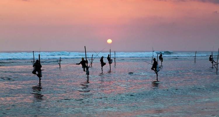 Silhouetten van traditionele paalvissers in evenwicht boven zachte oceaangolven bij zonsondergang met een roze lucht en ondergaande zon.