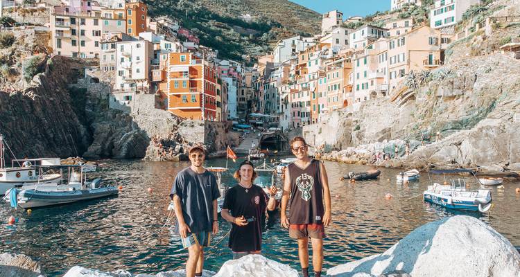 Trois amis prenant la pose sur des rochers devant le village coloré de Riomaggiore perché sur la falaise, Cinque Terre, avec de petits bateaux dans le port.