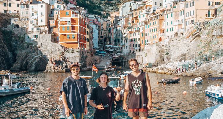Three travellers smiling at the waterfront of a colourful Cinque Terre village with boats on calm water