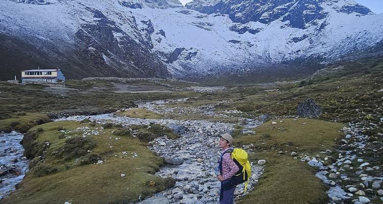 Randonneur solitaire se tient près d'un ruisseau bordé de pierres dans une large vallée himalayenne avec des plaques de neige