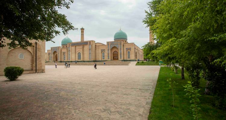 Cour spacieuse menant à un complexe de mosquée au dôme turquoise encadré de verdure par une journée nuageuse