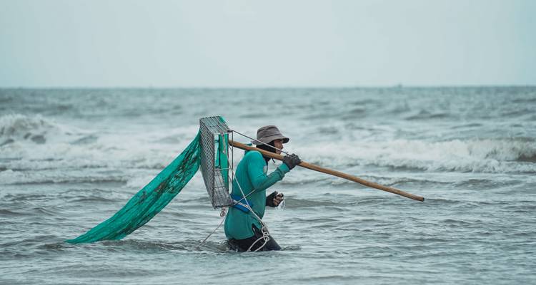 Visser waadt door oceaanbranding terwijl hij groene netconstructie meetrekt onder dreigende bewolkte hemel.