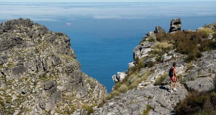 Solo hiker traverses rocky Table Mountain trail with deep blue Atlantic visible below the cliffs.