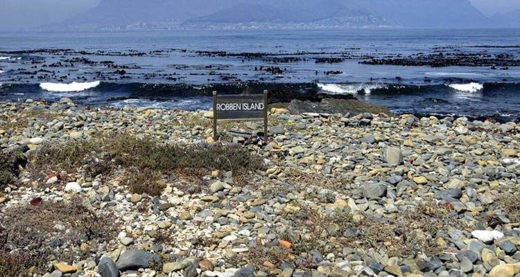 Rocky shoreline of Robben Island with a simple wooden sign and hazy Table Mountain in the background.