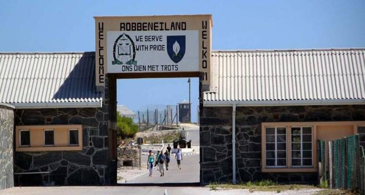 Entrance gate to Robben Island museum with visitors walking under bilingual welcome signs.