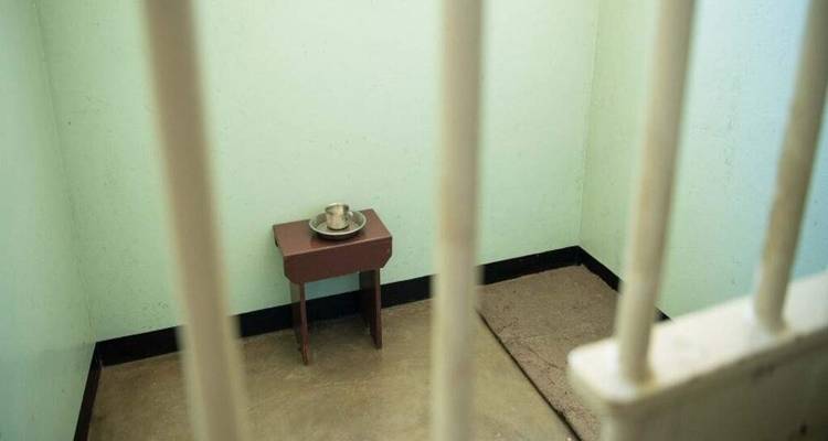 Simple prison cell with pale walls, a small table and tin cup viewed through metal bars