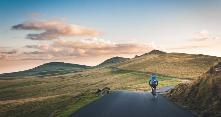 Fietser rijdend over een kronkelende asfaltweg door glooiende hooglandweiden onder een pastelkleurige avondhemel.