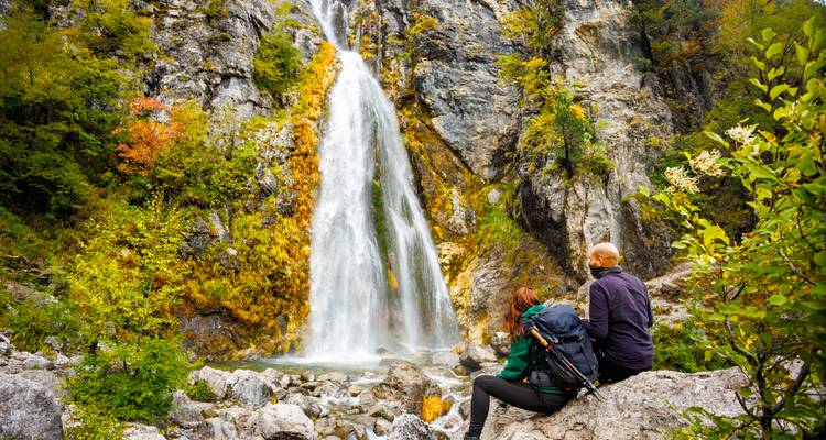 Twee wandelaars met rugzakken rusten uit op rotsen terwijl ze een hoge cascadewaterval bewonderen omgeven door herfstgebladerte.