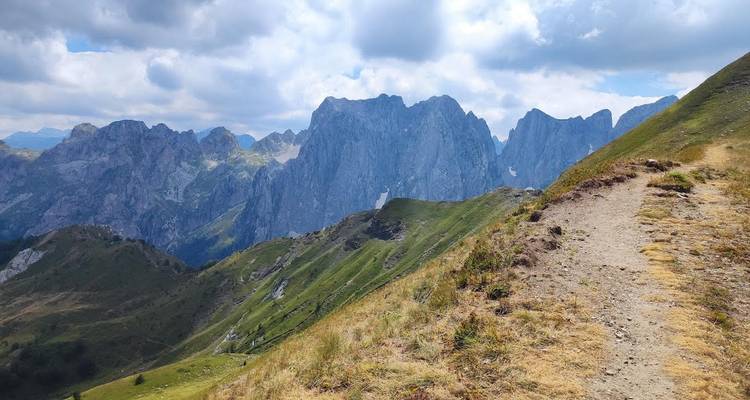 Sentier de montagne longeant une pente herbeuse escarpée avec des pics déchiquetés en arrière-plan