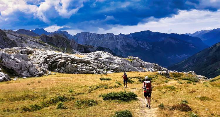 Randonneurs traversant une selle alpine herbeuse vers des pics escarpés sous des nuages dramatiques