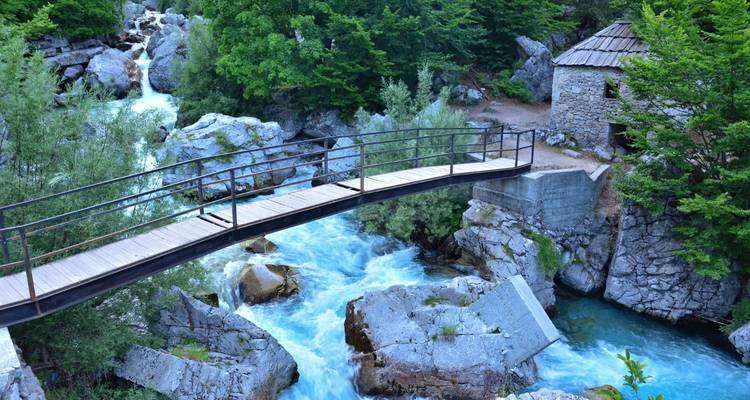 Étroit pont en bois enjambant un torrent de montagne bleu impétueux à côté d'une cabane en pierre
