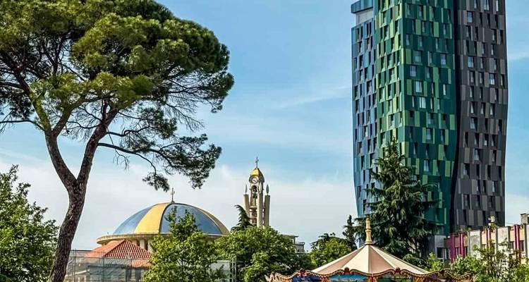 Rascacielos moderno de cristal que se eleva sobre un carrusel colorido y una mezquita con cúpula en el paisaje urbano de Tirana.
