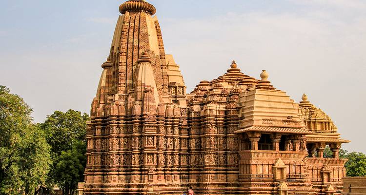 Un temple hindou de Khajuraho en grès richement sculpté se dresse contre un ciel pâle et des arbres verts.