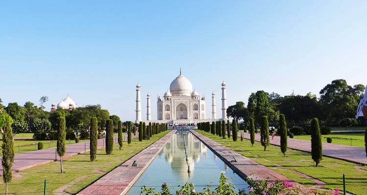 Vue frontale emblématique du Taj Mahal se reflétant dans le long bassin encadré par des jardins à la française sous un ciel bleu dégagé.