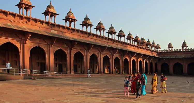 Des groupes de locaux en tenues colorées traversent la cour ensoleillée d'un complexe de mosquée en grès rouge.