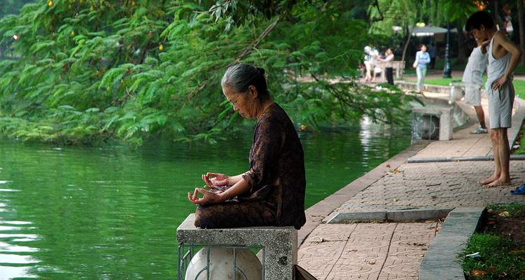 Ältere Frau meditiert neben grünem Seewasser, während Menschen in der Nähe in Hanoi Sport treiben.