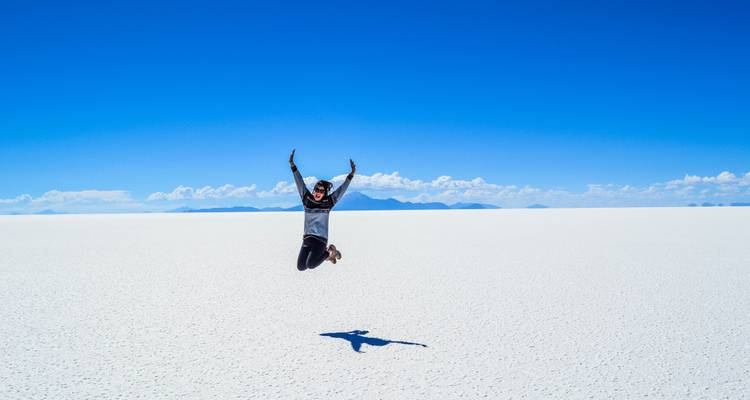 Voyageur en plein saut au-dessus de la surface blanche infinie du Salar d'Uyuni sous un ciel bleu éclatant, montagnes lointaines à l'horizon.