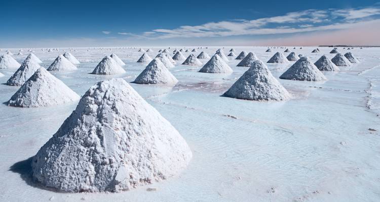 Des tas coniques bien ordonnés de sel récolté se reflétant sur l'eau peu profonde du Salar d'Uyuni sous un ciel bleu dégagé.