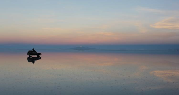 Silueta de un vehículo 4x4 y un viajero solitario en el salar de Uyuni como un espejo bajo la luz pastel del amanecer.