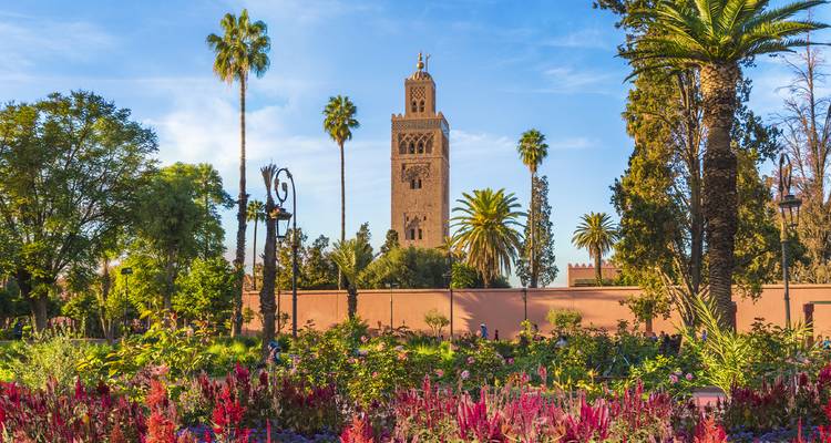 El alto minarete de la Mezquita Kutubía de Marrakech se alza sobre exuberantes jardines de flores y palmeras contra un cielo despejado.