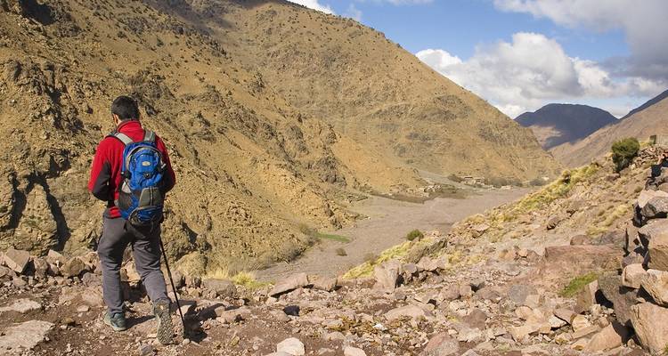 Excursionista solitario con chaqueta roja camina por senderos rocosos del Alto Atlas con bastón de senderismo entre montañas escarpadas color canela.