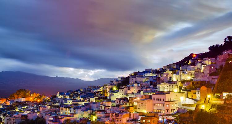 Chefchaouens blau gestrichene Hanghäuser, die unter dramatischen Abenddämmerwolken und Bergkulisse leuchten