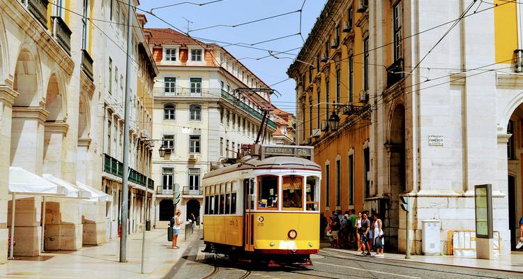 Classic yellow tram navigating a narrow historic street in Lisbon flanked by pastel buildings and cafés