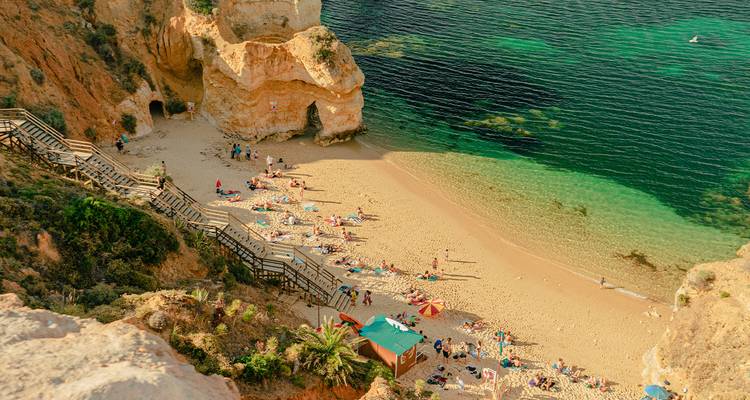 Scenic golden sand cove fringed by cliffs and emerald water with sunbathers and umbrellas seen from above.