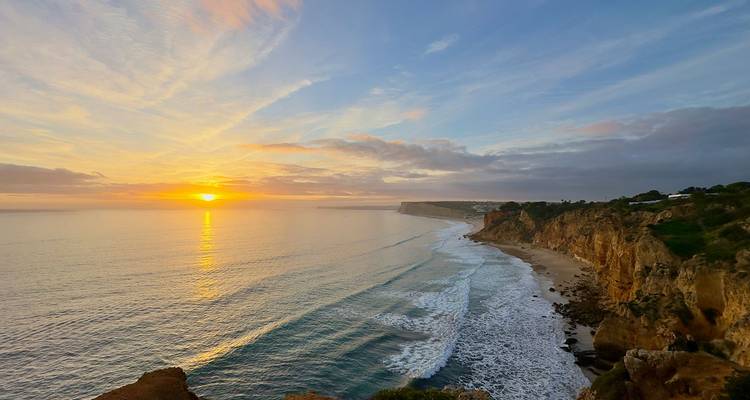 Sonnenuntergang mit warmem Licht über dramatischen Atlantikklippen und einem langen, weiten Strand an der Algarve