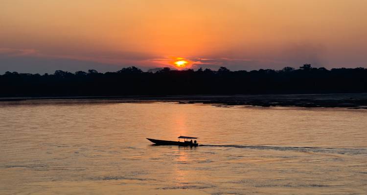 Pequeña lancha motora silueteada en un amplio río selvático durante una vívida puesta de sol naranja cerca de Puerto Maldonado.