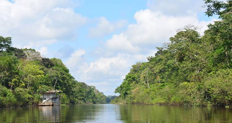 Corredor tranquilo de río en la selva con una pequeña casa flotante navegando entre orillas cubiertas de densa vegetación verde bajo un cielo parcialmente nublado.