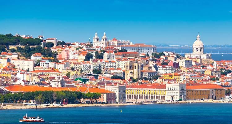 Panoramic skyline of Lisbon with white domes, red-tiled roofs and Tagus River in foreground.