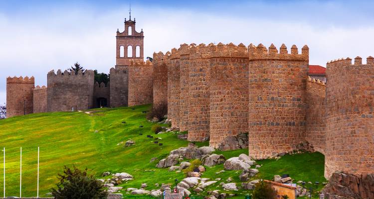 Massieve middeleeuwse stenen muren en kantelen torens van Ávila die oprijzen boven een grasachtige helling en rotspartijen.