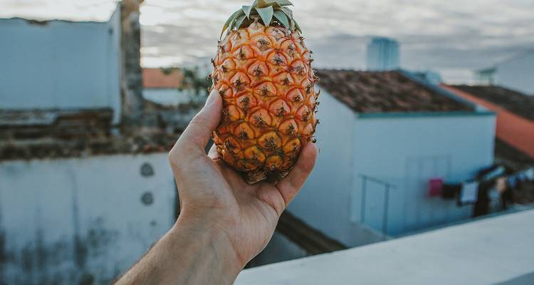Close-up van een hand die een kleine rijpe ananas vasthoudt tegen een achtergrond van betegelde daken en een zachte avondlucht.