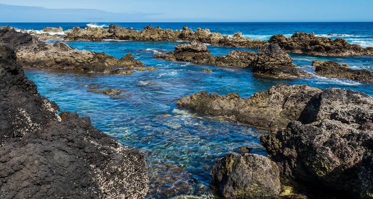 Jagged black lava rocks enclose crystal-clear tidal pools beside a deep blue ocean.