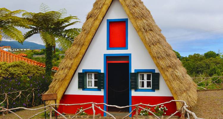 Maison traditionnelle triangulaire au toit de chaume avec des accents rouge vif, bleu et blanc entourée de fleurs à Madère.