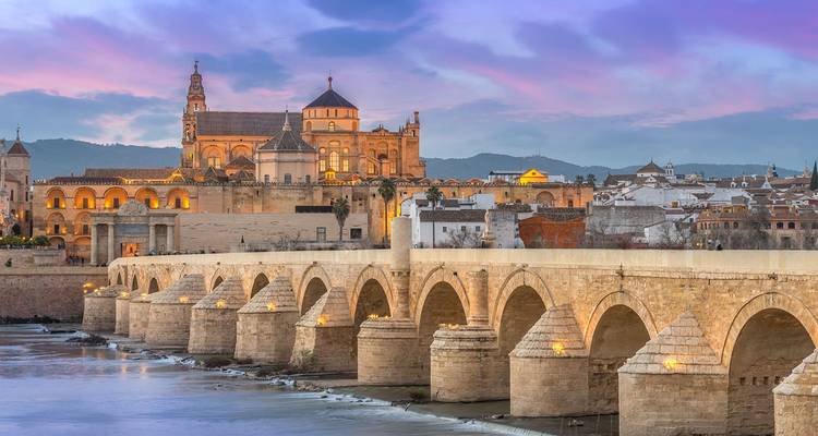 Die Römische Brücke und die beleuchtete Moschee-Kathedrale von Córdoba in der Abenddämmerung unter einem farbenfrohen Himmel.