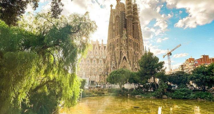 La basílica de la Sagrada Família resplandeciendo bajo la luz del sol vespertino detrás de un estanque y exuberante vegetación