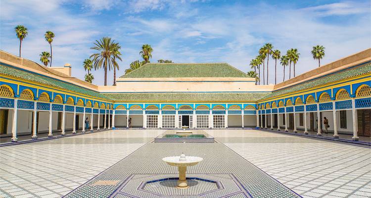 Ruime betegelde binnenplaats met kleurrijke Moorse bogen, centrale fontein en met palmen omzoomde daklijn onder een heldere blauwe hemel.