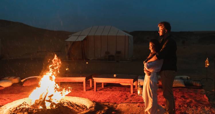 Jong stel knuffelend naast een laaie kampvuur in een woestijnkamp bij schemering, met silhouetten van canvas tenten achter hen.