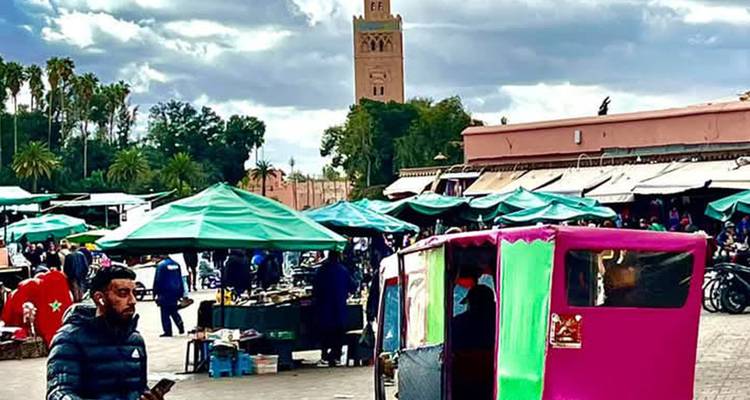 Drukke marktplein met kraampjes, kleurrijke riksja en de Koutoubia-moskeetoren die op de achtergrond oprijst.