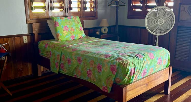 Eenvoudig houten eenpersoonsbed met kleurrijk bloemenbeddengoed in een rustieke huttenkamer met luiken en een staande ventilator.