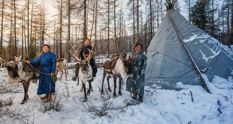 Pastores de renos de pie con sus animales junto a un tipi en un bosque nevado