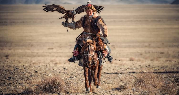 Cazador de águilas tradicional con vestimenta de piel montando a caballo a través de la estepa mientras sostiene un águila dorada en alto.