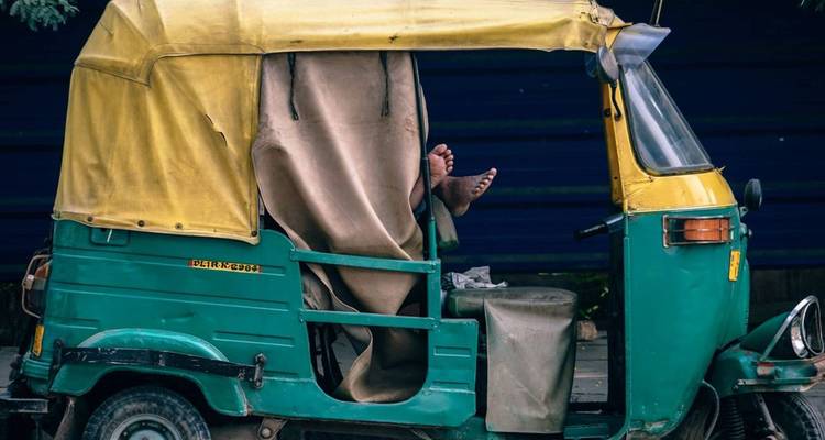 Auto-Rikscha mit herausragenden Füßen des Fahrers, geparkt auf einer indischen Straße.