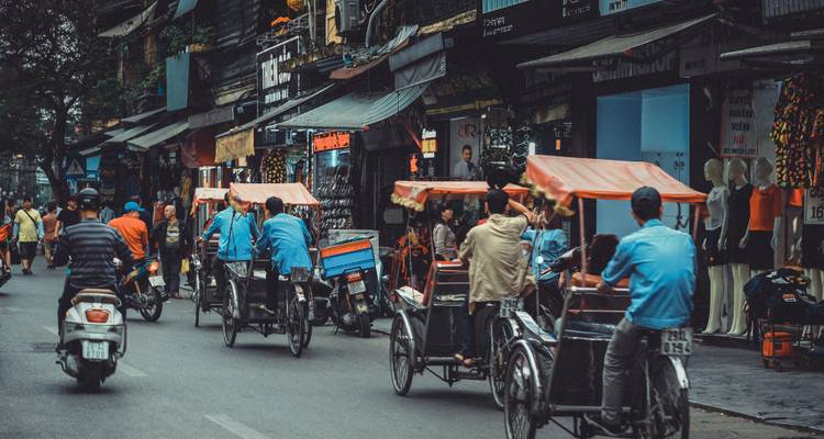 Rue animée de la vieille ville avec des cyclo-pousses et des scooters qui zigzaguent devant les devantures de magasins à Hanoï.